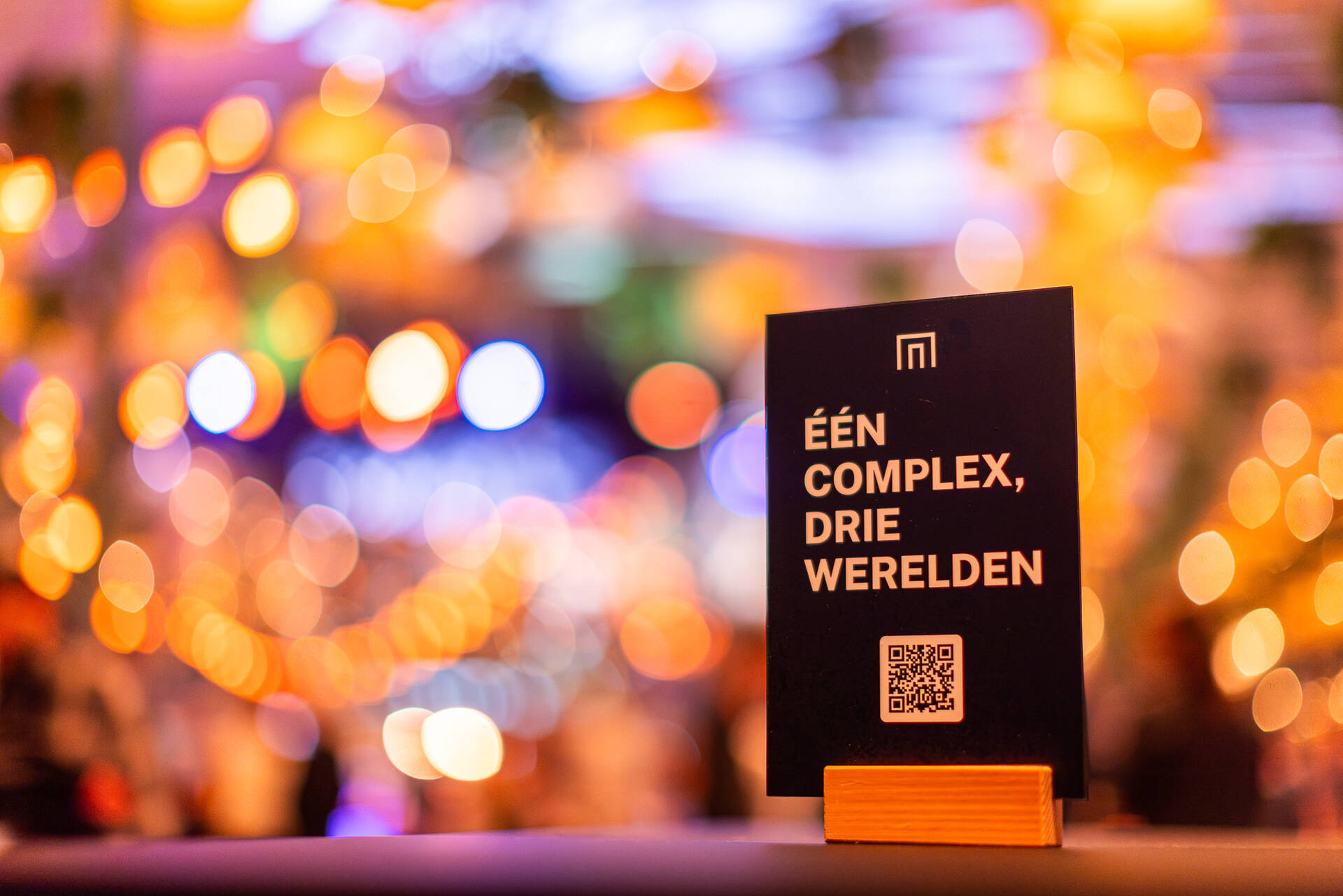 Een bordje met de tekst "Een complex, drie werelden" en lichtjes wazig op de achtergrond